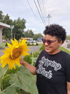 a light skinned black woman with curly hair wearing glasses and a black shirt with gray letters  that read "Dripping in Black" smiles wide at a large sunflower